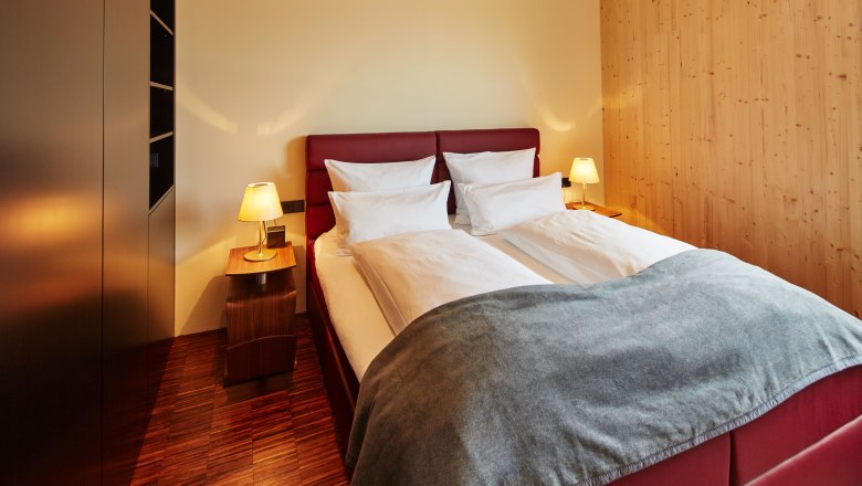 Spálňa v podkroví záhrady Guntrams, © Herbert Lehmann Útulná spálňa s manželskou posteľou, červeným čelom, dvoma nočnými stolíkmi s lampami a drevenými stenami.