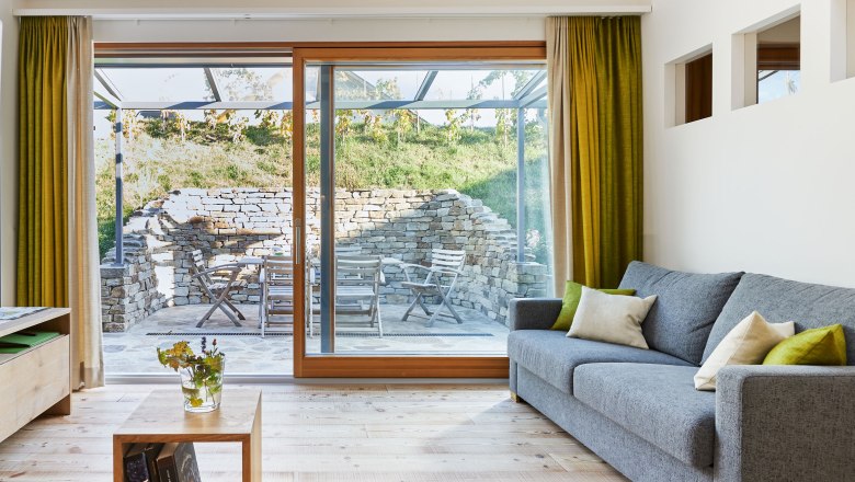 Weinviertler Kellerstöckl, © Andreas Hofer Pohľad do interiéru modernej obývacej izby so sivou pohovkou, drevenou podlahou a veľkými oknami vedúcimi na terasu s kamennou stenou a záhradným nábytkom.