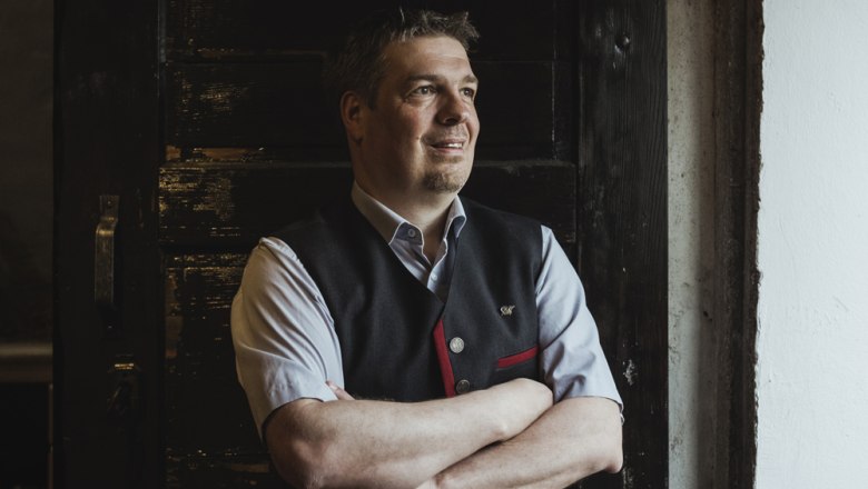 Prenajímateľ Dietmar Schöner, © Niederösterreich Werbung/David Schreiber Muž v tradičnom odeve stojí so založenými rukami pred tmavými drevenými dverami.