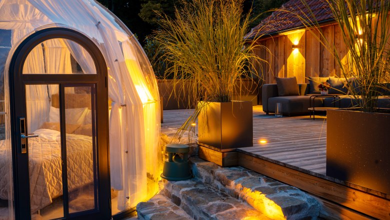 Chaty a miesta plné ZÁZRAKOV - Weidehaus De Luxe, © WUNDERVOLL eben Moderná chata s terasou a priehľadným kupolovitým stanom v lese za súmraku.