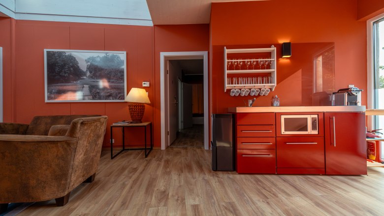 Chaty Grafenegg, © Niederösterreich Werbung / Maximilian Pawlikowsky Pohľad do interiéru modernej obývačky s červenými stenami, červenou kuchynskou linkou, hnedými kreslami a veľkým oknom.