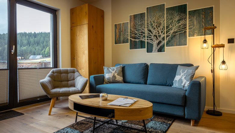 Ötscher Lodge, © Niederösterreich Werbung / Maximilian Pawlikowsky Útulná obývacia izba s modrou pohovkou, kreslom, dreveným stolom a výhľadom na les cez okno.