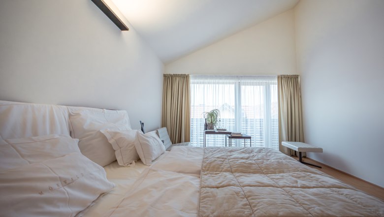 MÖRWALD, Relais Chateaux Hotel am Wagram, © Niederösterreich Werbung / Maximilian Pawlikowsky Moderná hotelová izba s veľkou posteľou, bielymi vankúšmi a prehozmi, svetlými závesmi a oknom v pozadí.