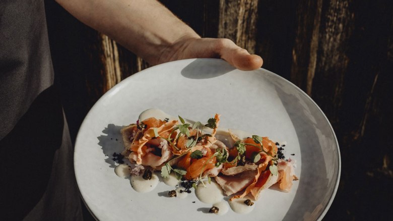 Vitello Forello, © Niederösterreich Werbung/Sophie Menegaldo Jednou rukou sa podáva tanier umne naaranžovaného vitello forello, ozdobený bylinkami a omáčkou.