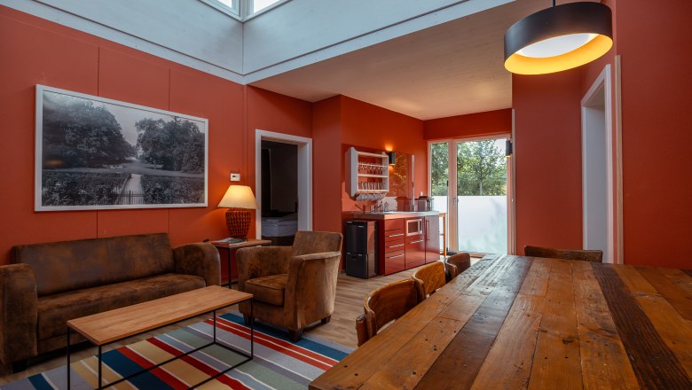 Chaty Grafenegg, © Niederösterreich Werbung / Maximilian Pawlikowsky Pohľad do interiéru modernej obývačky s červenými stenami, dreveným stolom, pohovkou a kuchynským kútom.