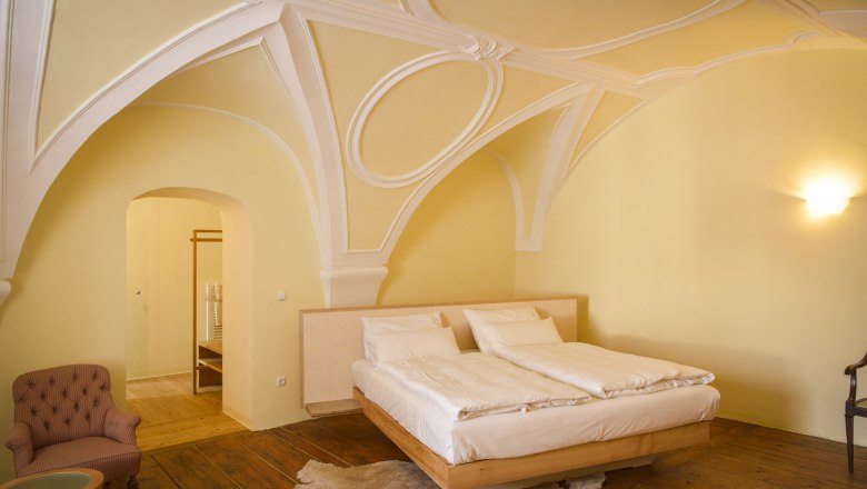 Oberstockstall Estate, © Robert Herbst Elegantná spálňa s klenutým stropom, manželskou posteľou, kreslom a drevenou podlahou.