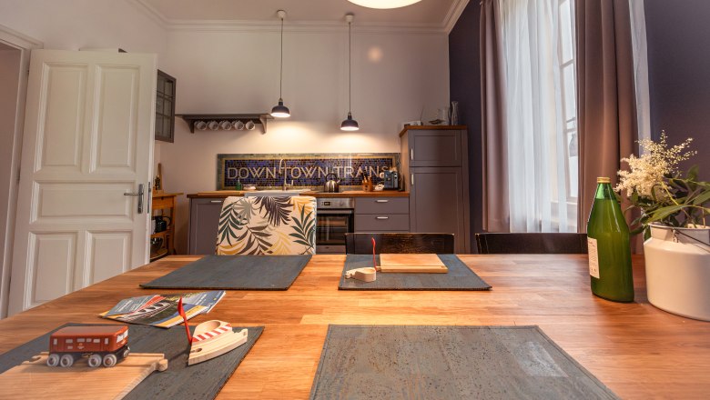 Dovolenka v hoteli BAHNhof, © Niederösterreich Werbung / Maximilian Pawlikowsky Kuchyňa s dreveným stolom, hračkárskym vlakom, fľašou a kvetmi na stole, modernými lampami a kuchynskými skrinkami v pozadí.