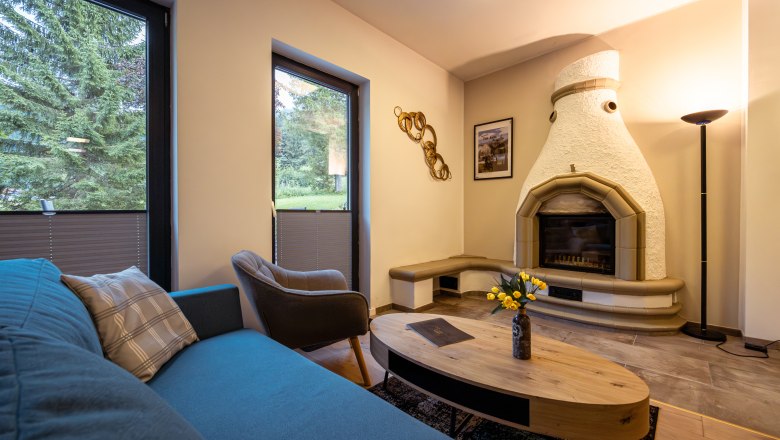 Ötscher Lodge, © Niederösterreich Werbung / Maximilian Pawlikowsky Útulná obývacia izba s krbom, pohovkou, kreslom a konferenčným stolíkom. Veľké okná s výhľadom na zeleň.