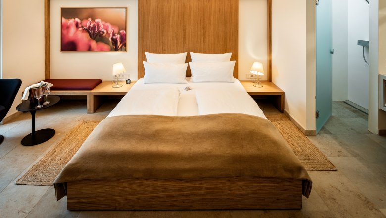 Penzión Flora, © Herbert Lehmann Moderne zariadená hotelová izba s manželskou posteľou, nočnými stolíkmi, lampami a obrazom na stene.