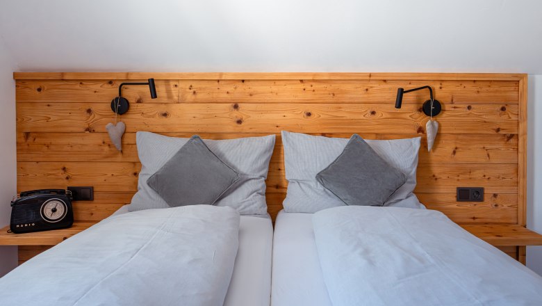 Leopold an der Ybbs, © © Niederösterreich Werbung/ Maximilian Pawlikowsky Útulná manželská posteľ s dreveným operadlom, sivými vankúšmi a prikrývkami. Naľavo retro rádio, napravo lampa na čítanie a dekorácia srdca.