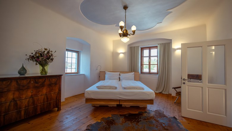 Oberstockstall Estate, © Niederösterreich Werbung / Maximilian Pawlikowsky Útulná spálňa s drevenou podlahou, manželskou posteľou, starožitnou komodou a kyticou kvetov. Stropné svetlo a okno so závesmi.