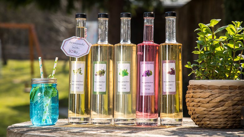 Domáce sirupy z bylinkovej záhrady hostinca Rath-Smetana, © Niederösterreich Werbung/Kurt Pinter Päť fliaš domáceho sirupu a modrý pohár so slamkami na drevenom stole vonku.