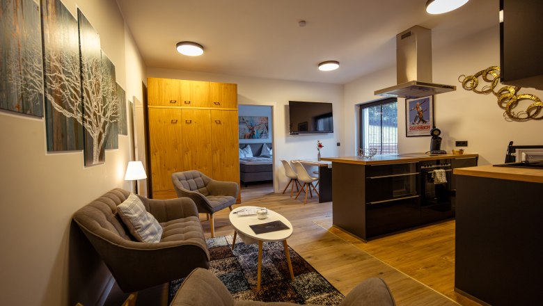 Ötscher Lodge, © Niederösterreich Werbung / Maximilian Pawlikowsky Moderná obývacia izba s kuchyňou, pohovkou, kreslami a jedálenským stolom. Na stenách je vidieť svetlý drevený nábytok a umelecké diela.