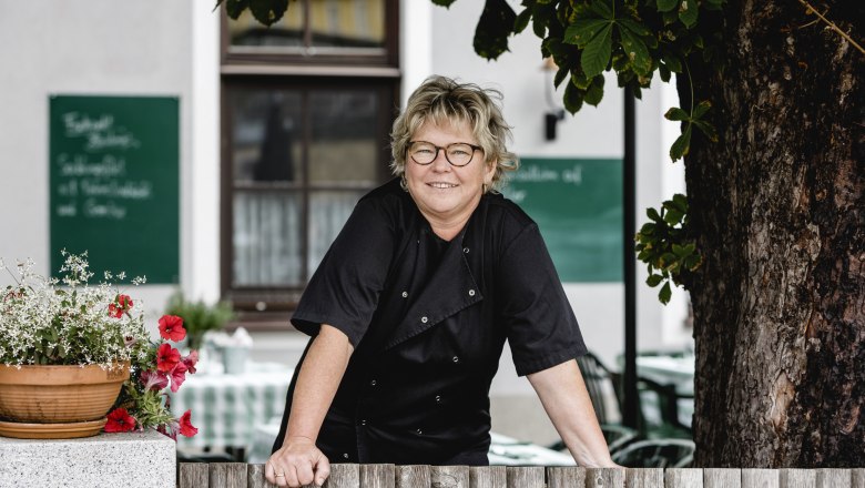 Domáca pani Martina Ebner, © Niederösterreich Werbung/David Schreiber Žena v čiernom sa opiera o drevený plot, v pozadí sú kvety a strom.