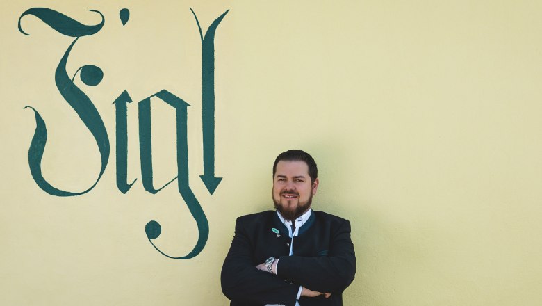 Matthias Strunz, prenajímateľ Figl, © Niederösterreich Werbung/David Schreiber Muž v tradičnom odeve stojí pred žltou stenou s nápisom Figl.