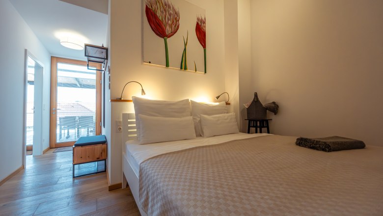Apartmány Hausgnost, © Niederösterreich Werbung / Maximilian Pawlikowsky Moderná spálňa s manželskou posteľou, svetlou drevenou podlahou a umeleckým dielom na stene. Dvere vedú na balkón s výhľadom na strechy.