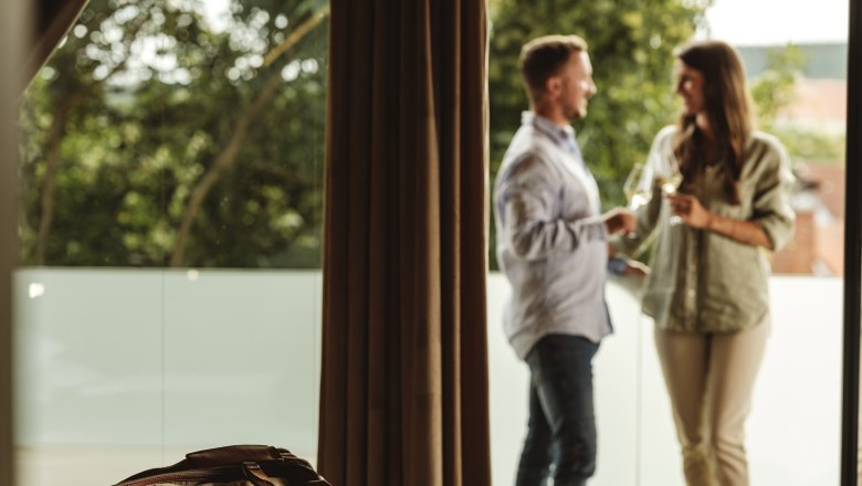 Vinárstvo Hagn Kellerstöckl, © Weinviertel Tourismus GmbH Pár stojí na balkóne a drží poháre s vínom, v popredí leží na posteli cestovná taška.