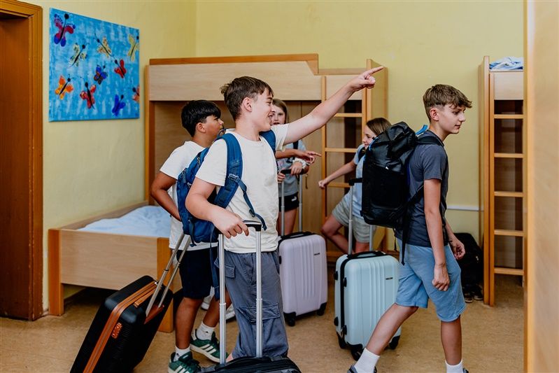 Skupina detí s kuframi v ubytovni s poschodovými posteľami.