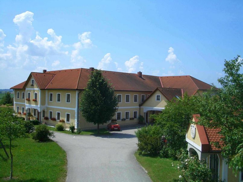 Veľká farma so žltými múrmi a červenými strechami, obklopená stromami a cestou.