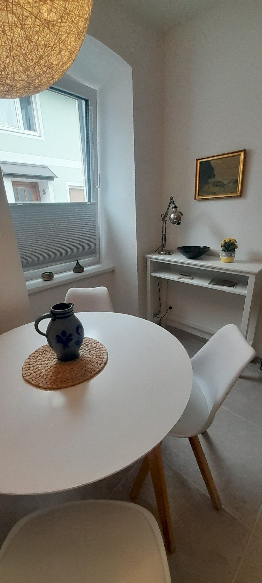 Malá jedáleň s okrúhlym bielym stolom, dvoma bielymi stoličkami a ozdobnou vázou. V pozadí okno a malý stolík s lampou.