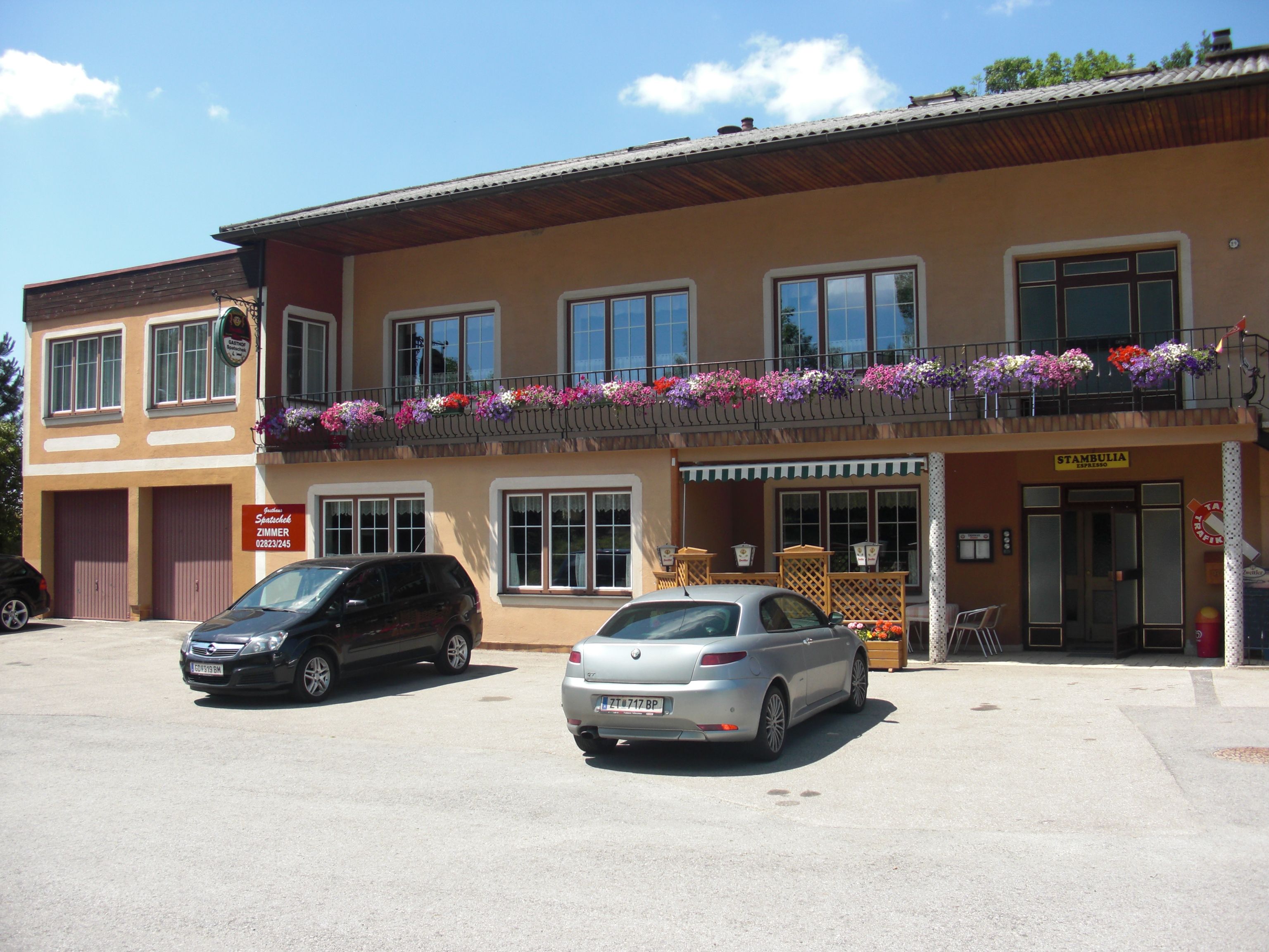 Dom pre hostí s balkónom a kvetmi, dve zaparkované autá vpredu.