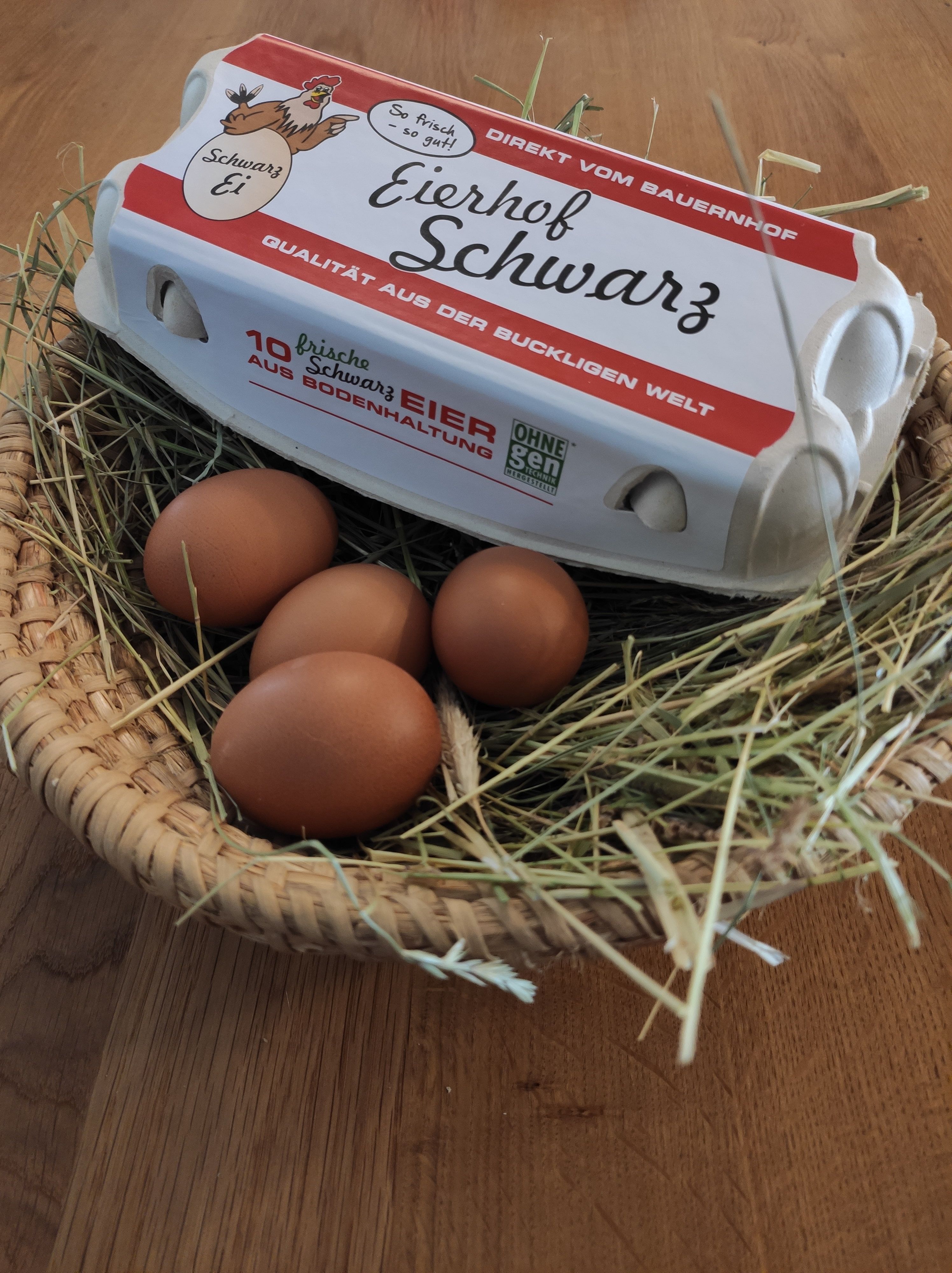 Kartón na vajcia s nápisom "Eierhof Schwarz" leží na košíku so slamou a hnedými vajcami.