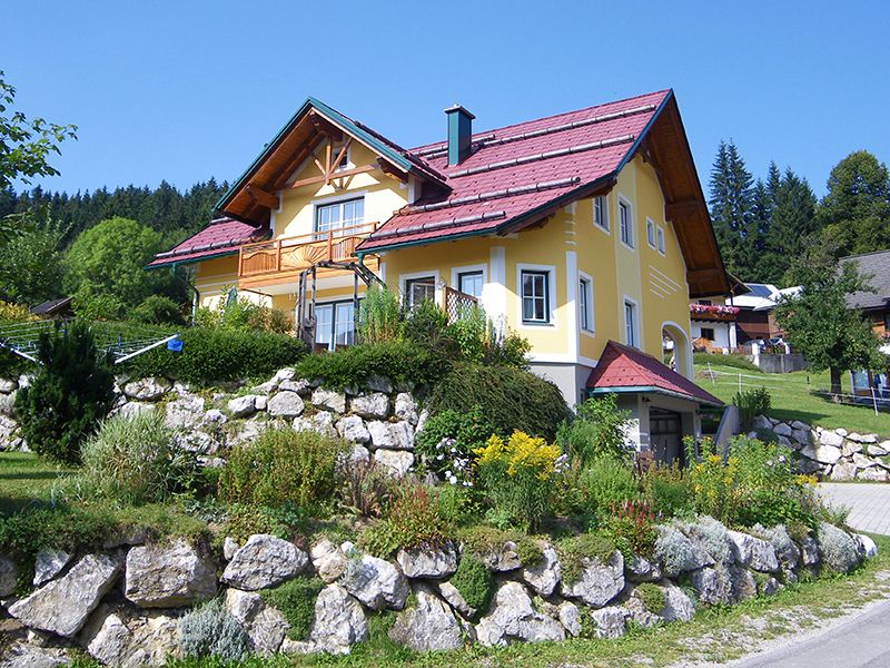 Žltý dom s červenou strechou v zelenej krajine.