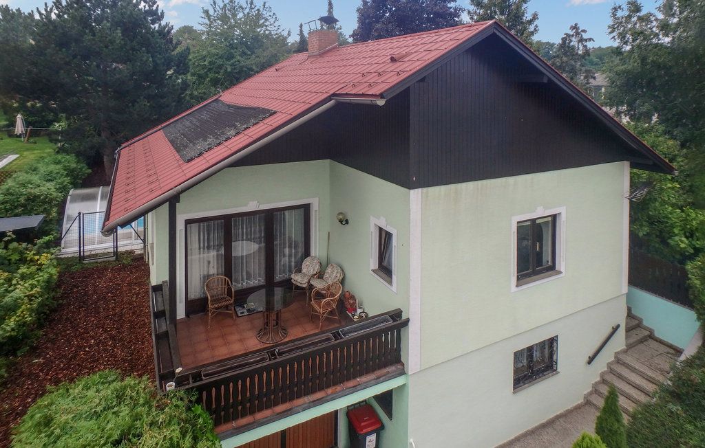 Dvojpodlažný dom s červenou strechou a balkónom, obklopený stromami.