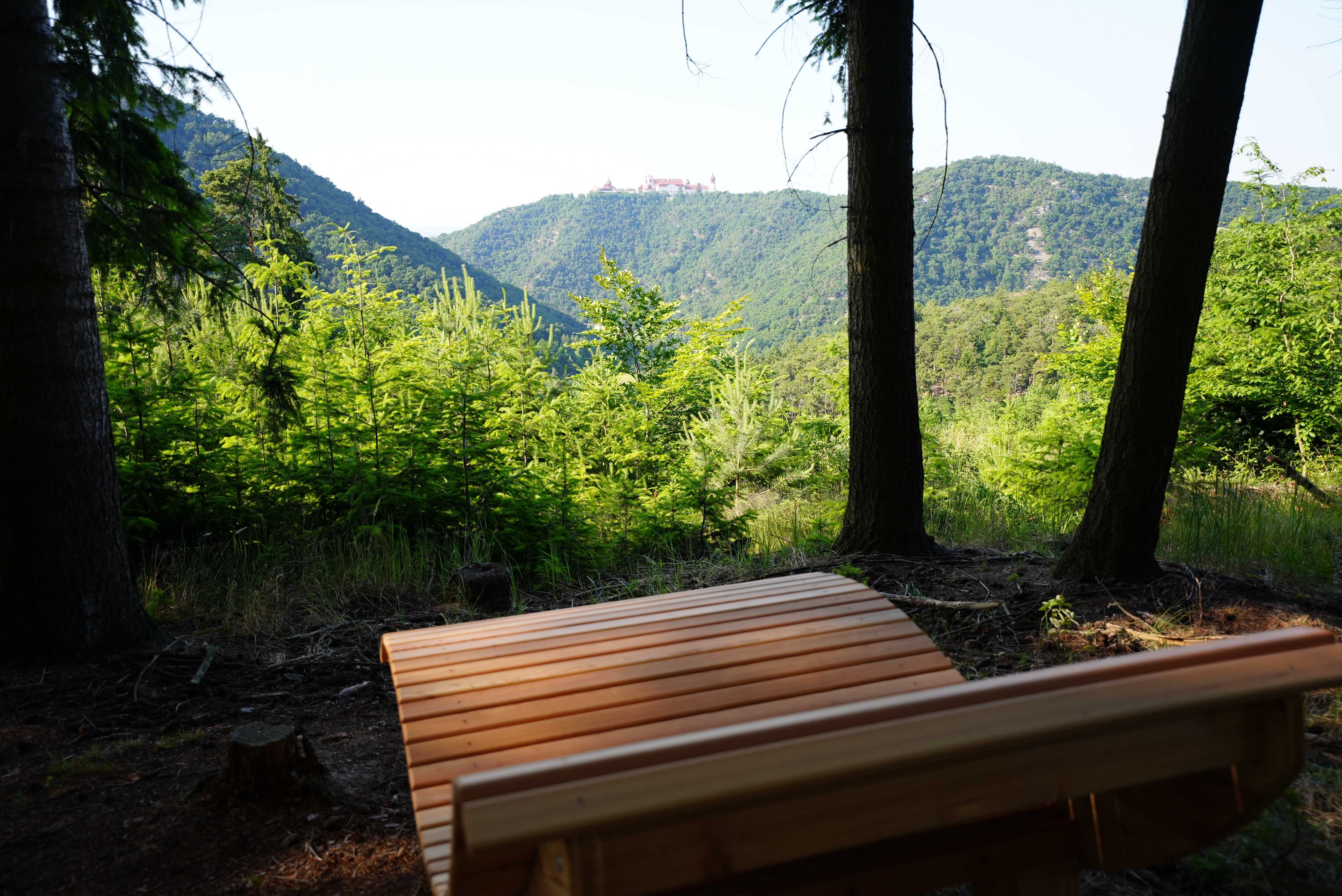 Drevená lavička v popredí s výhľadom na zalesnené kopce a kláštor v diaľke.