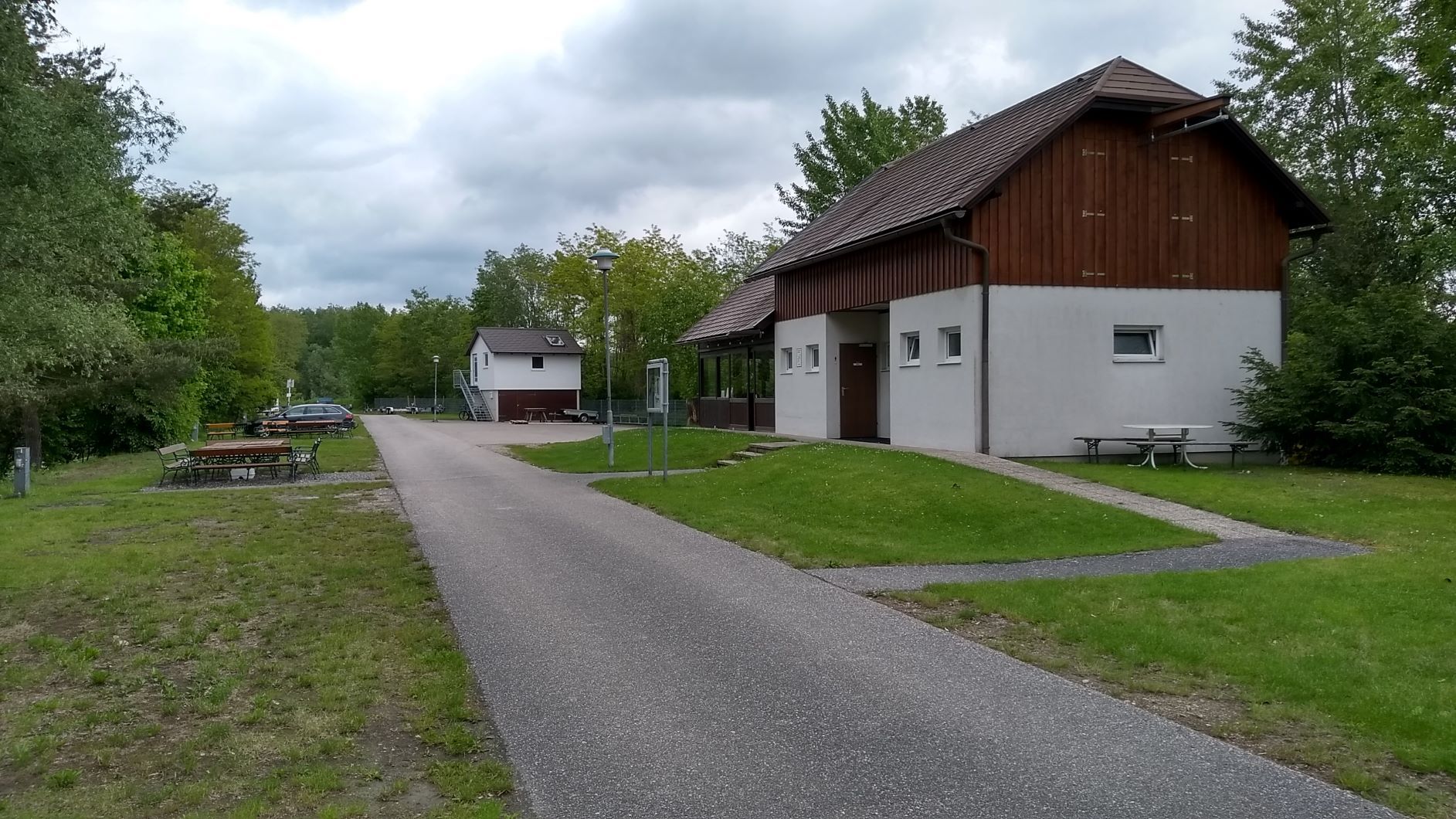 Cesta vedie okolo bielej budovy s dreveným obložením, obklopenej stromami a lúkami, pod zamračenou oblohou.