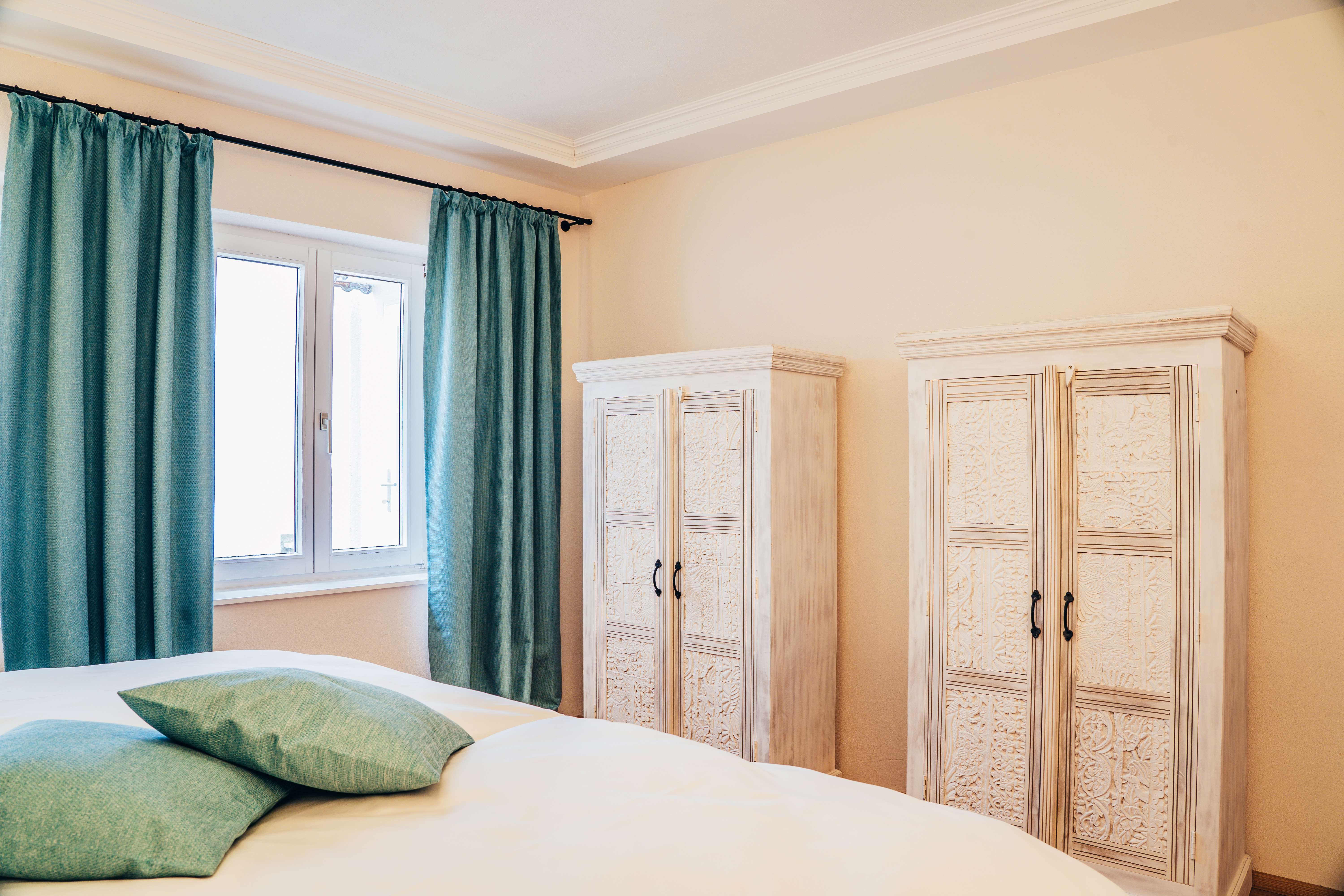 Spálňa s dvoma bielymi skriňami, tyrkysovými závesmi a vankúšmi.