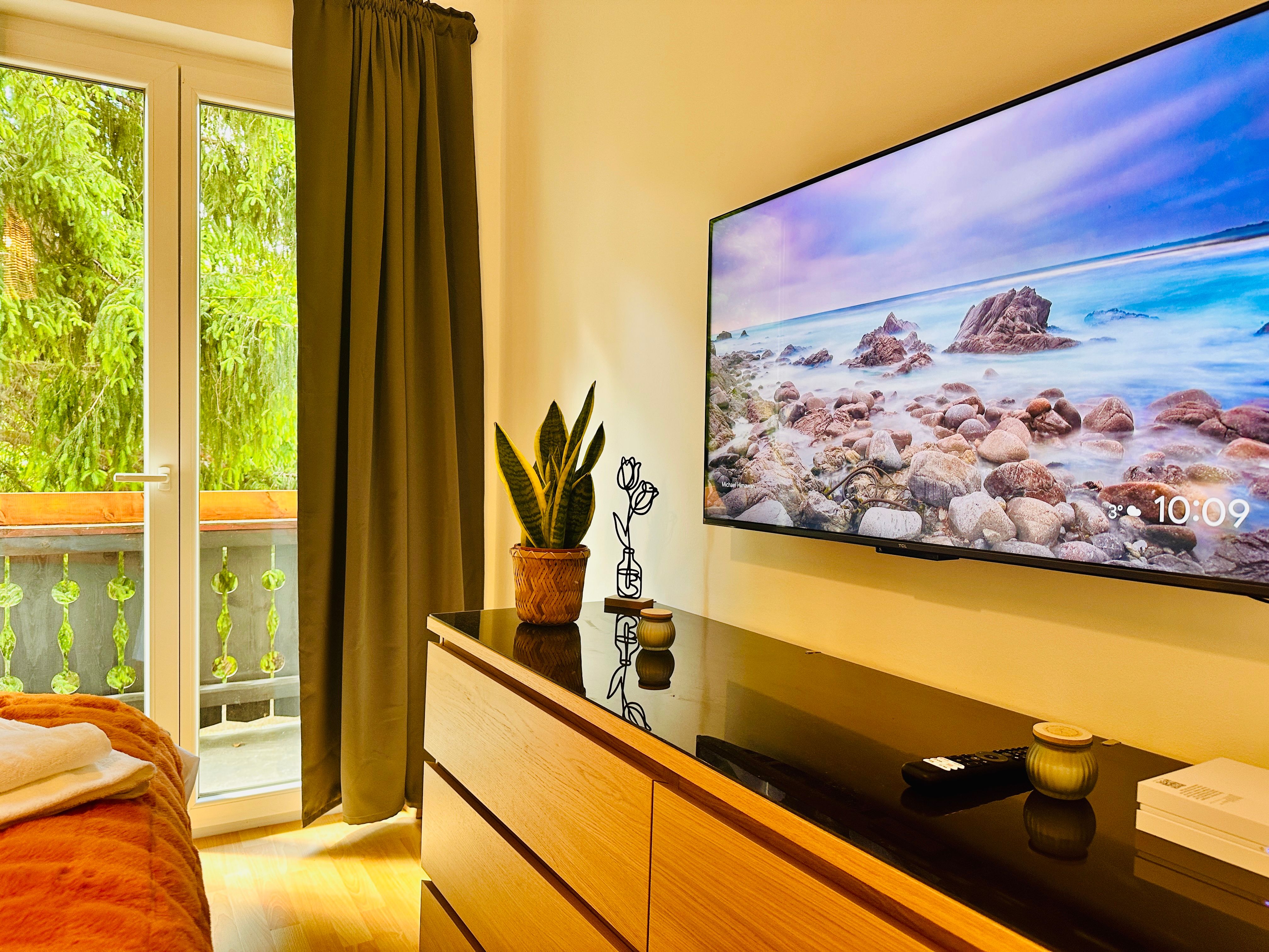 Útulná izba s TV, komodou, rastlinou a výhľadom na balkón.