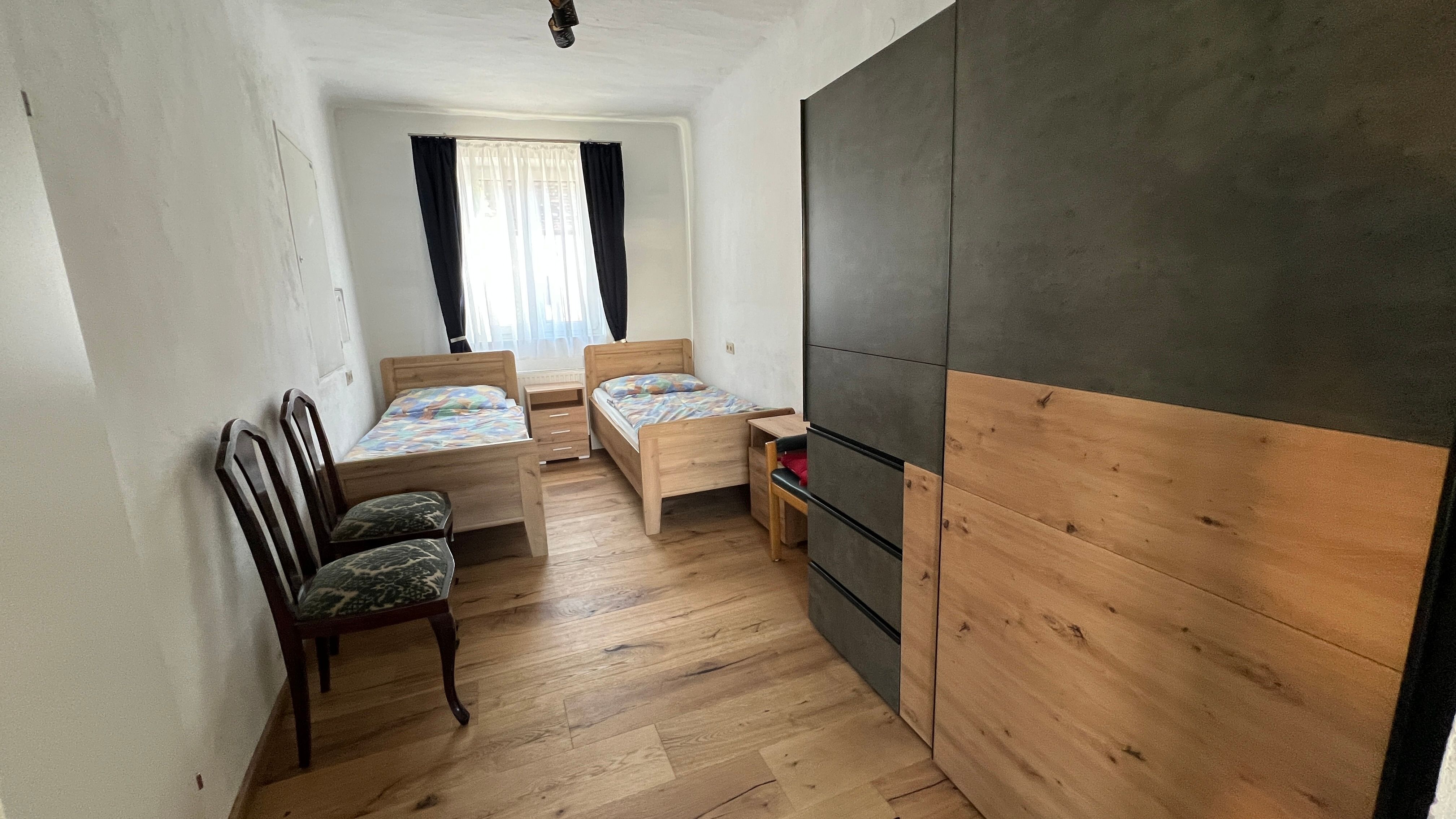 Malá spálňa s dvoma samostatnými posteľami, šatníkovou skriňou a dvoma stoličkami. Okno so závesmi prepúšťa svetlo.