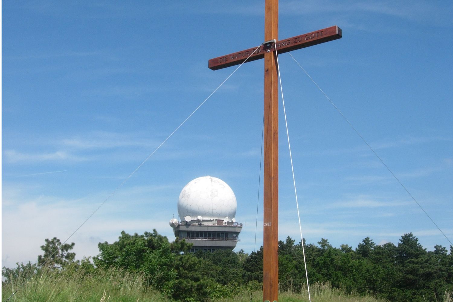 Drevený kríž pred radarovou kupolou na vrchu Buschberg, obklopený stromami a modrou oblohou.