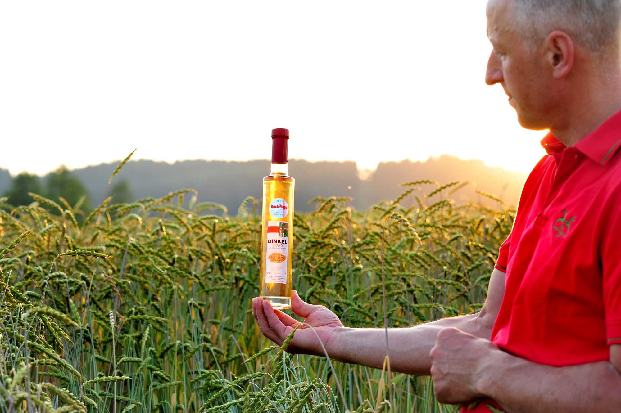 Muž v červenej košeli drží fľašu špaldovej pálenky na kukuričnom poli pri západe slnka.