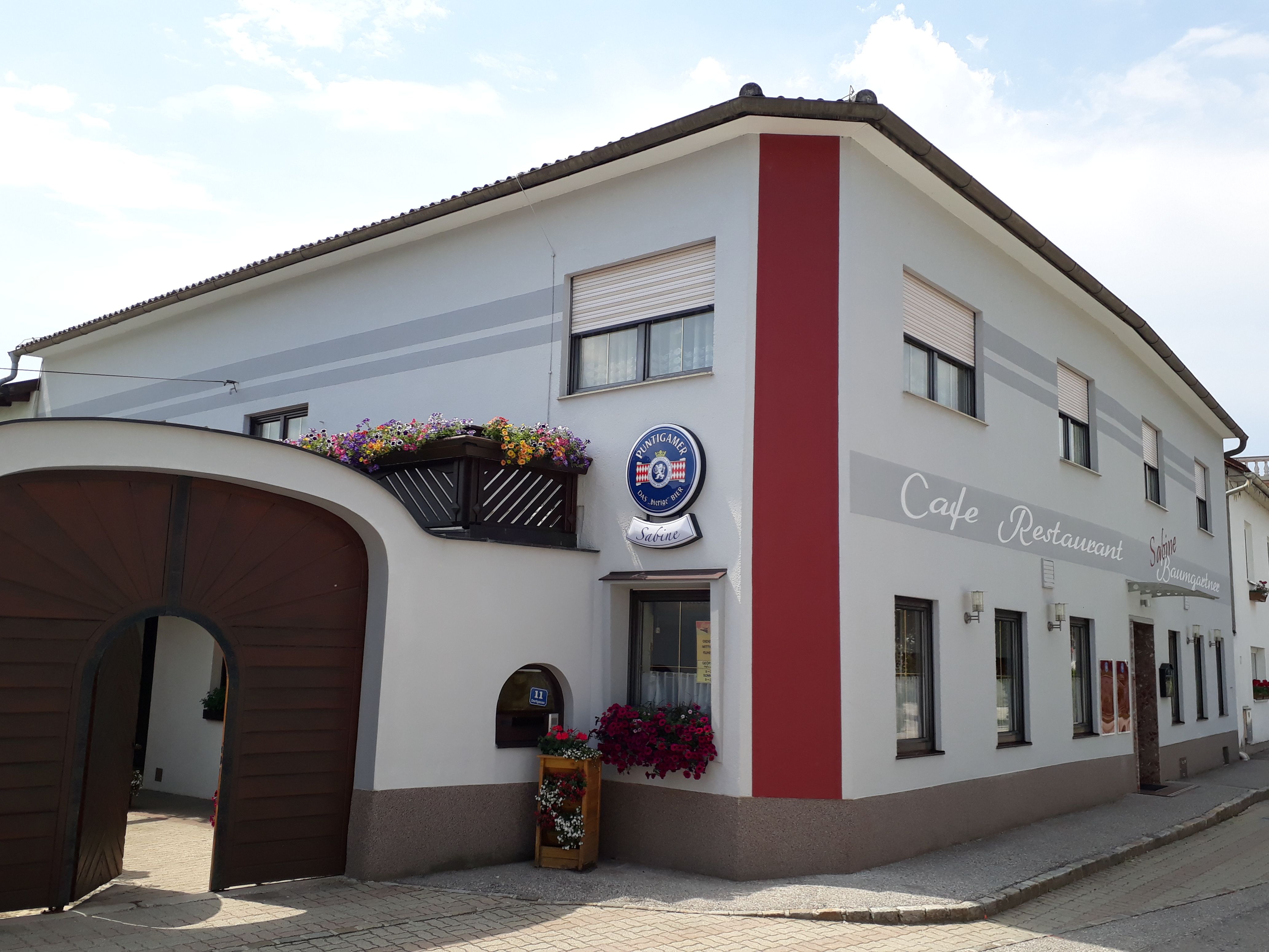 Exteriérový pohľad na kaviareň Café Sabine s drevenou bránou, bielo-červenou fasádou a nápisom "Café-Restaurant".