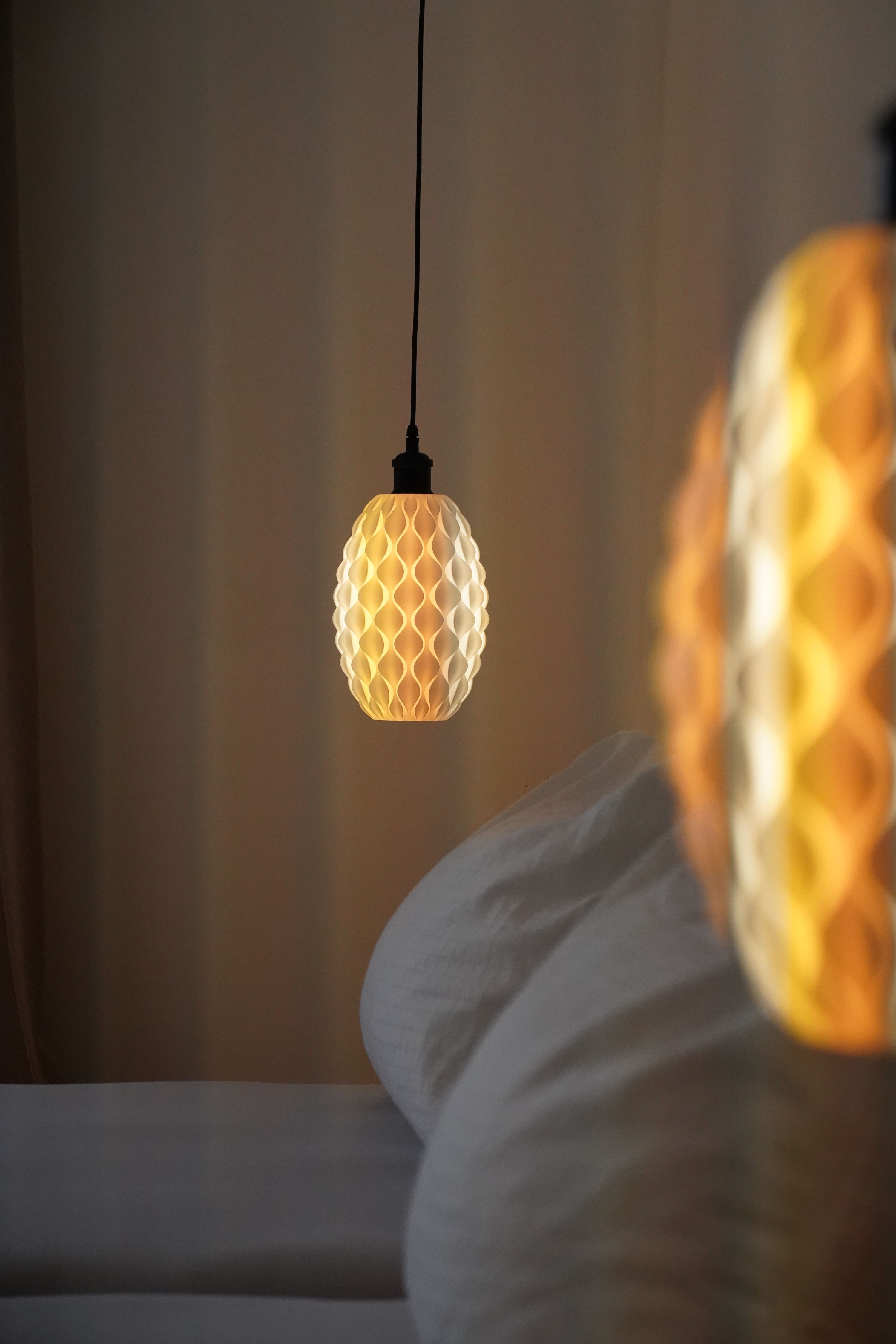 Detailný záber na závesnú lampu so vzorom včelích plástov, ktorá vyžaruje teplé svetlo vedľa postele.