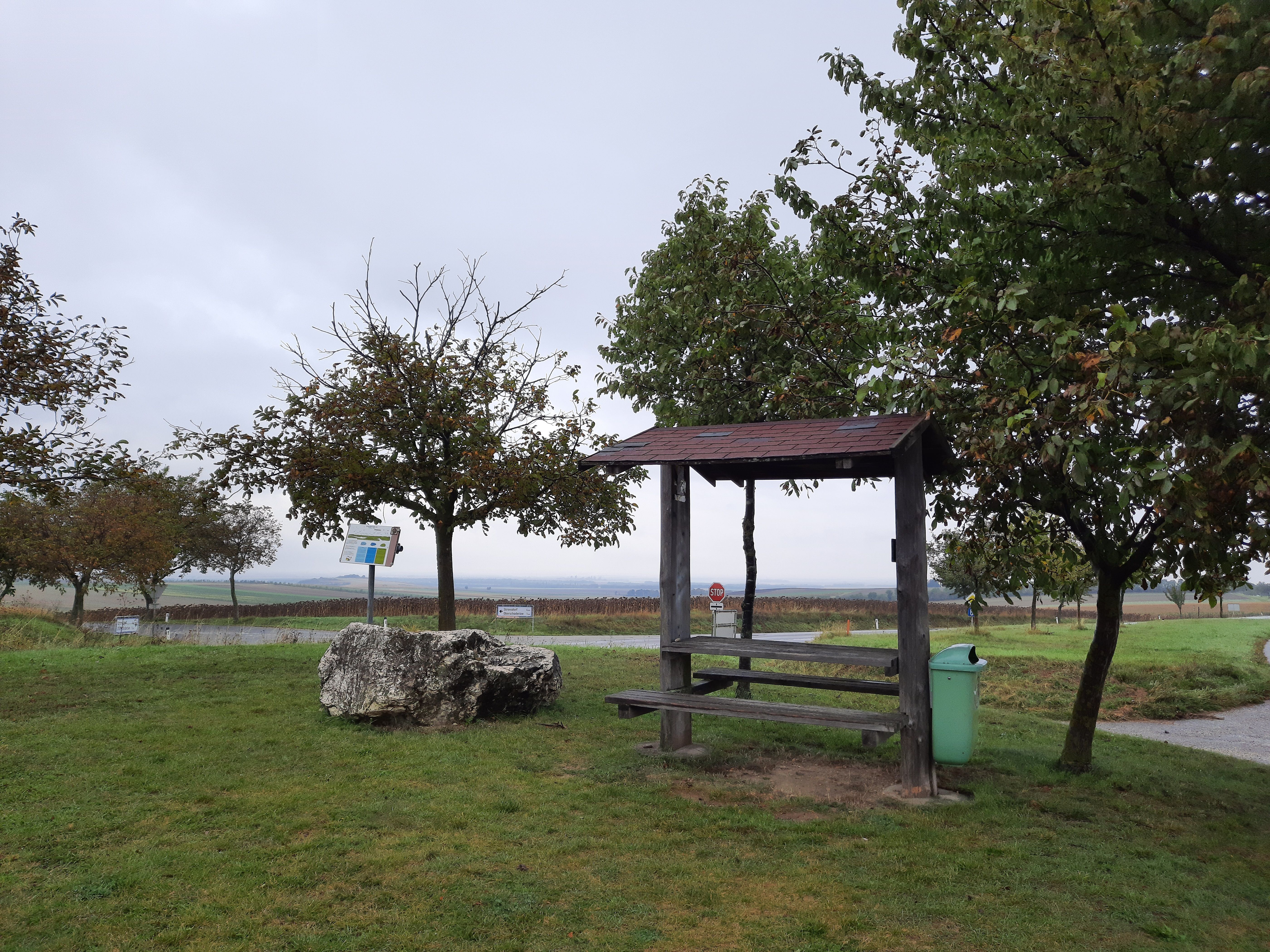 Odpočívadlo s drevenými lavicami a stolom, obklopené stromami a lúkou, pod zamračenou oblohou.