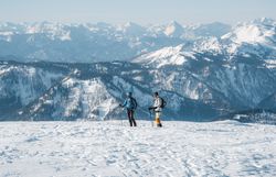 Die glitzernde Schneedecke erstreckt sich bis zum Horizont, während zwei Skitourengeher die unberührte Winterlandschaft erkunden. Die majestätischen Berge im Hintergrund bieten eine atemberaubende Kulisse und laden dazu ein, die Ruhe und Schönheit der Natur zu genießen.