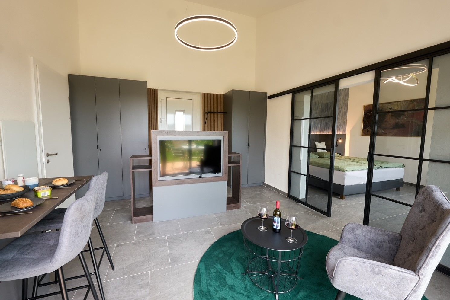Moderný byt s jedálenským stolom, TV, sivými skriňami a posuvnými sklenenými dverami do spálne.