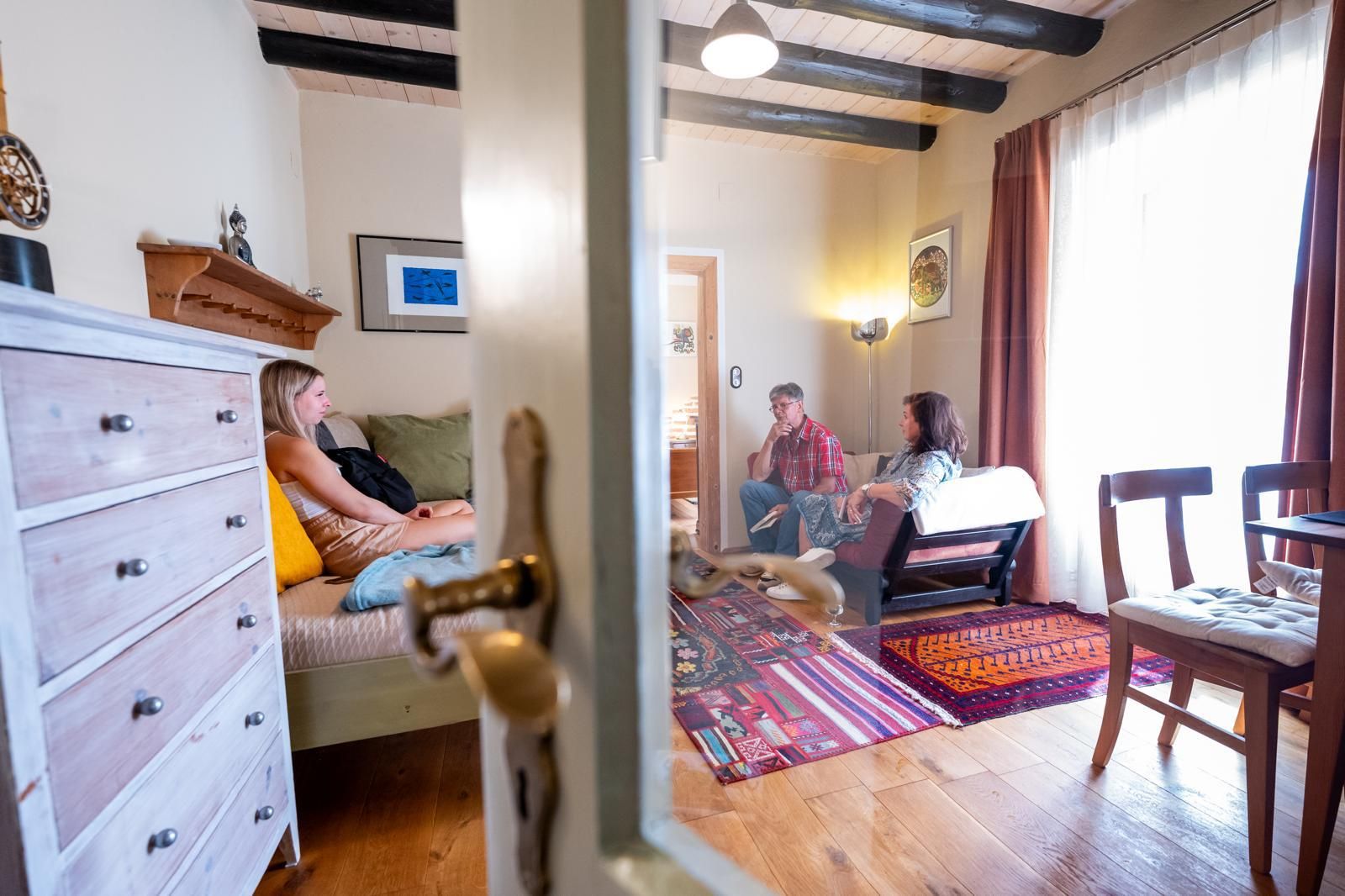 V útulnej obývačke s drevenou podlahou a farebným kobercom sedia tri osoby.