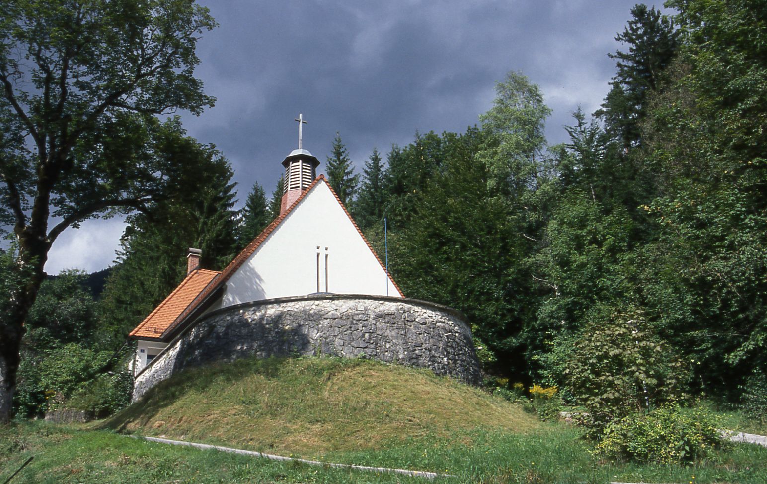 Malý kostolík v lese s kamenným základom a červenou strechou.