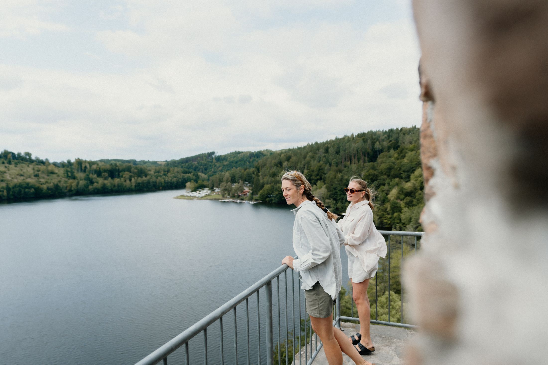 Dve ženy stoja na balkóne s výhľadom na jazero a zalesnené kopce.