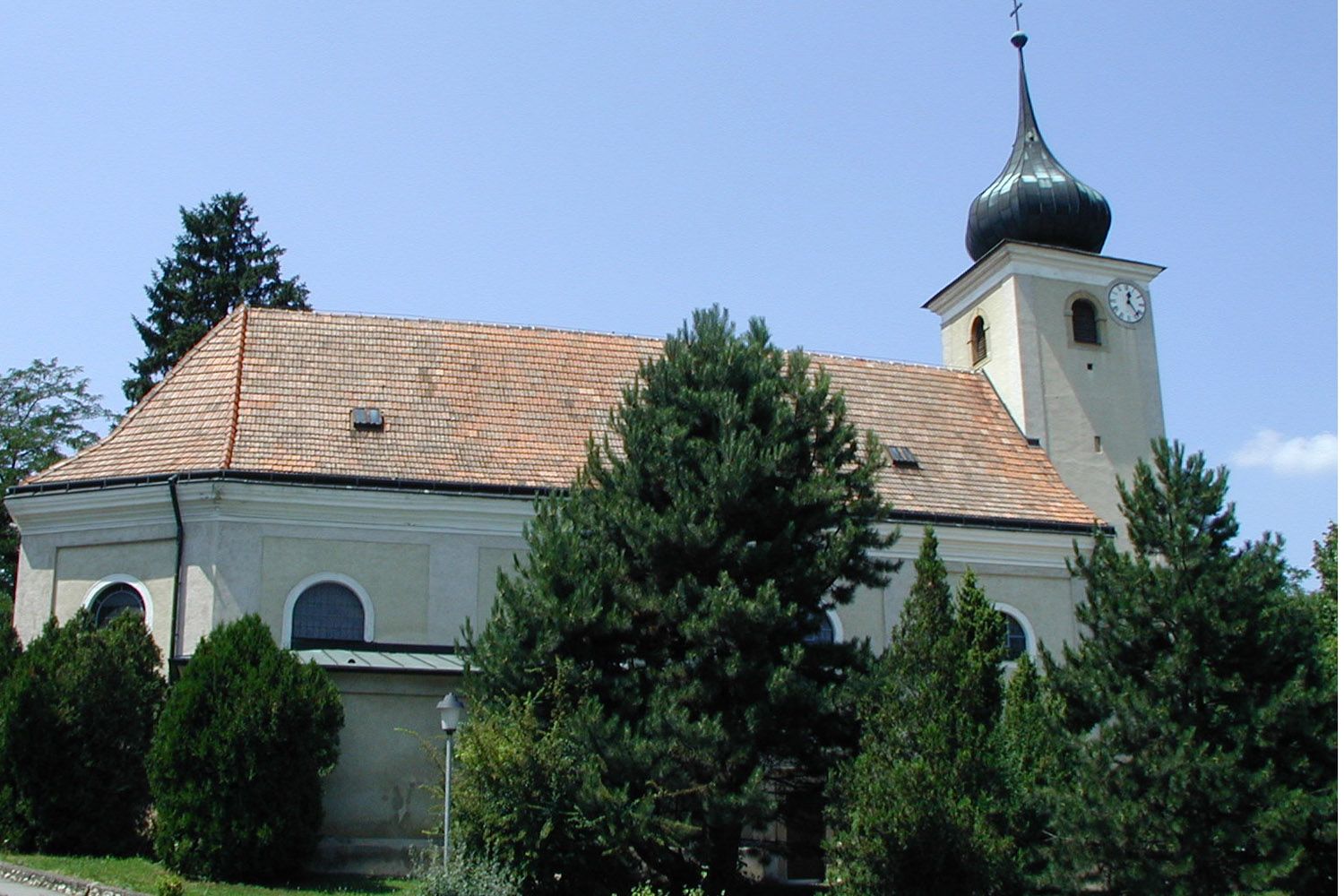 Kostol v Schleinbachu s vežou a hodinami, obklopený stromami.