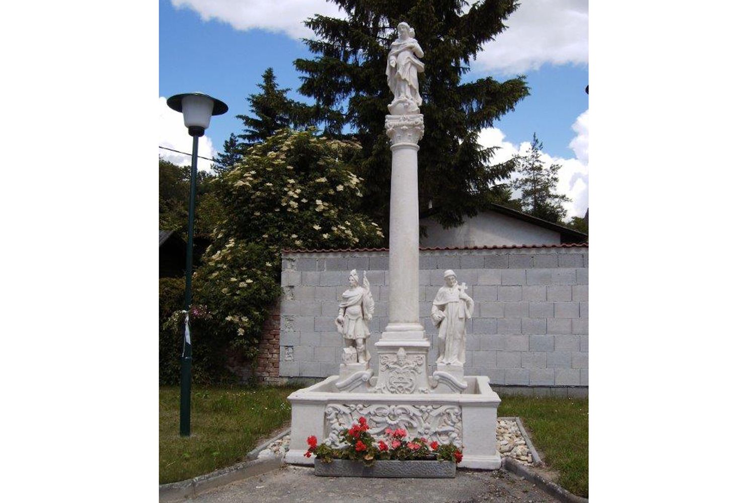 Mariánsky stĺp so sochami a kvetmi, obklopený stromami a múrom.