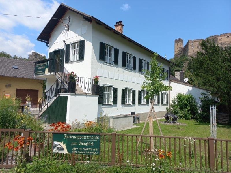 Biela budova so zelenými okenicami, obklopená záhradou, v pozadí hrad.