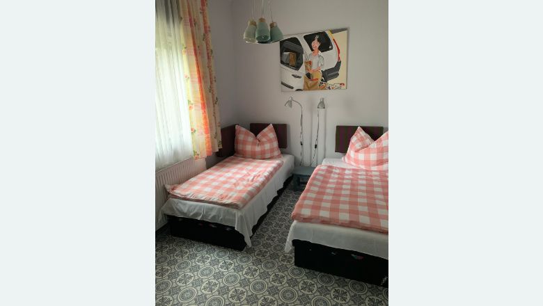Dve jednolôžkové postele s ružovými a bielymi šachovnicovými perinami v spálni so vzorovanou podlahou a obrazom na stene.