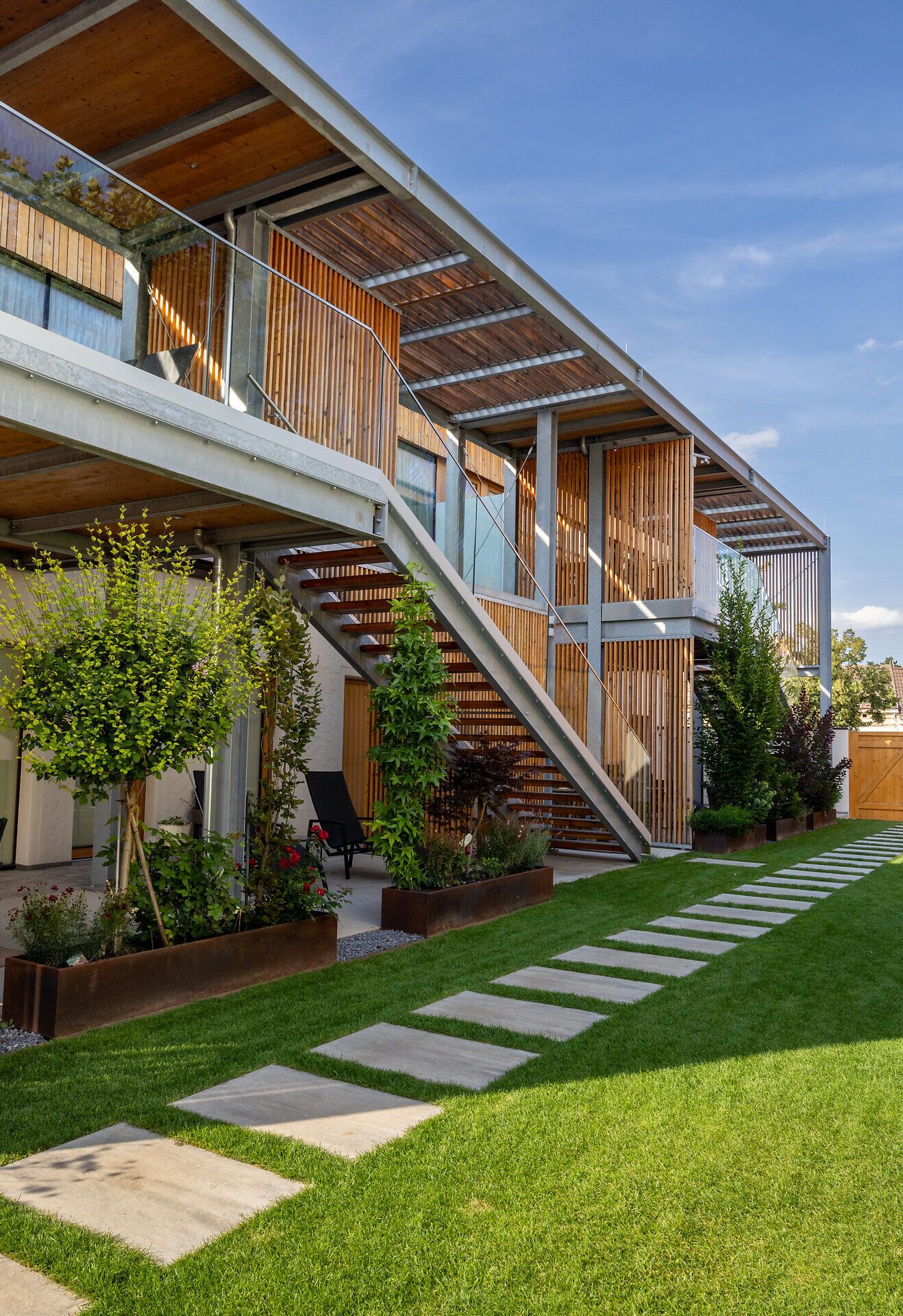 Moderná budova s bielou fasádou a drevenými prvkami, obklopená múrom s drevenou bránou.