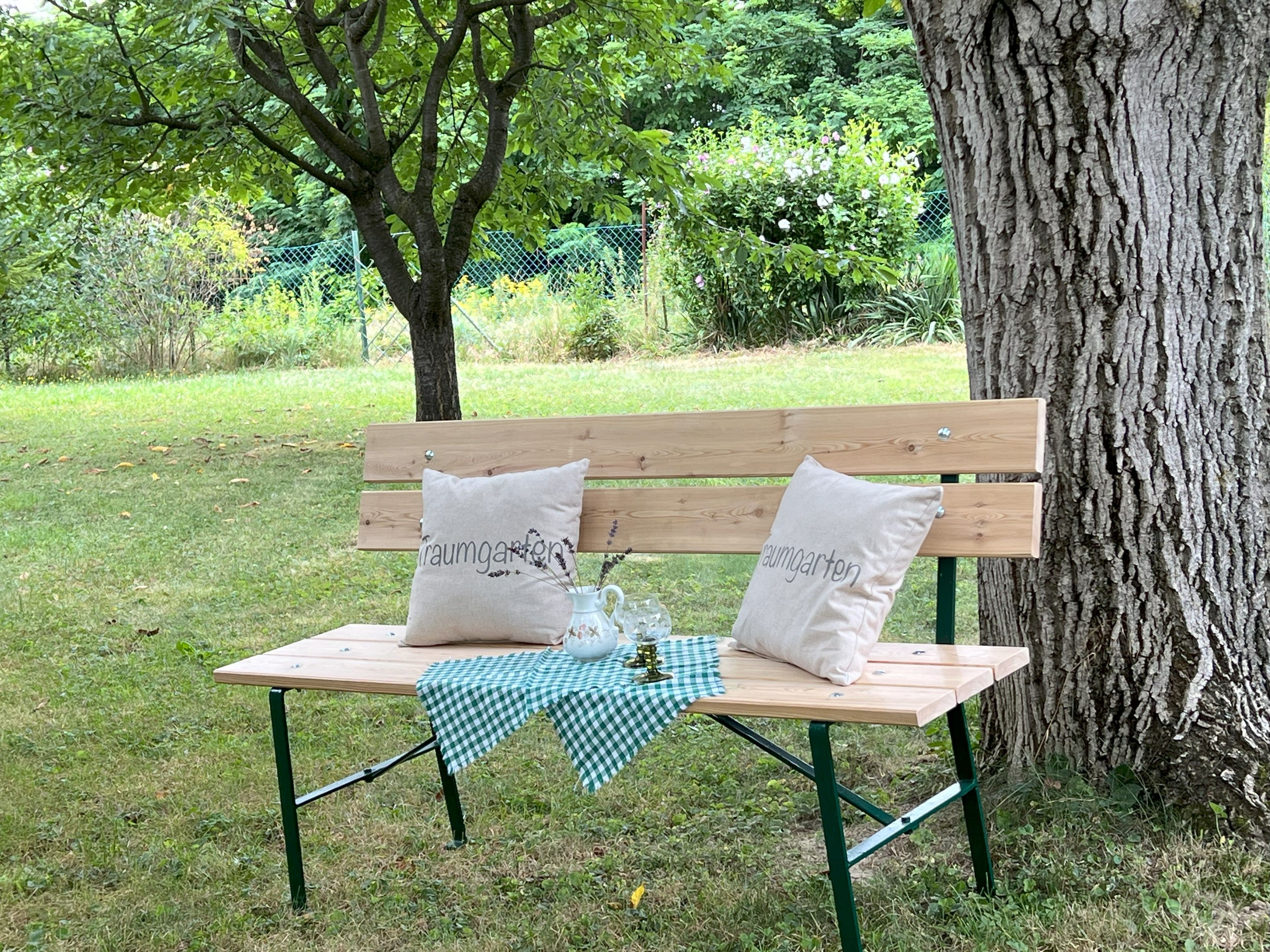 Drevená lavica s vankúšmi a obrusom pod stromom v záhrade.