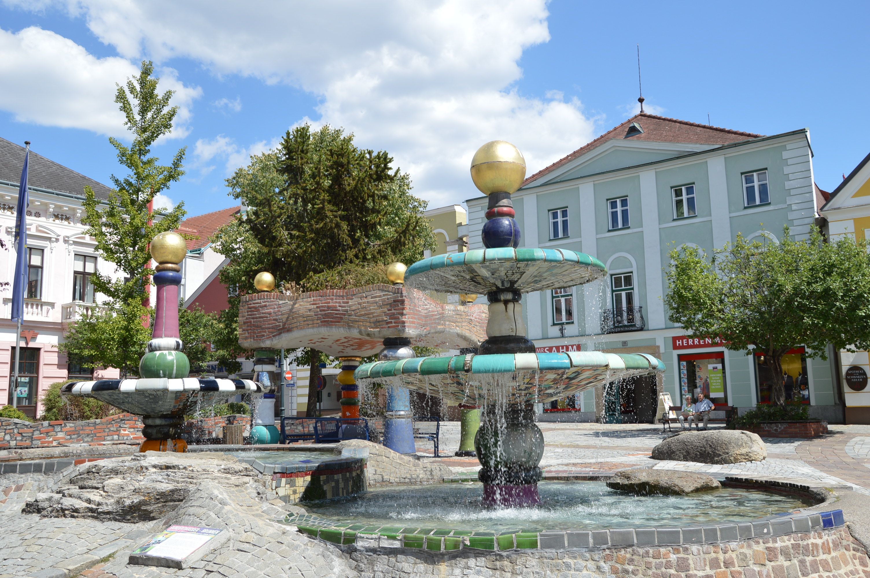 Farebná fontána v Hundertwasserovom štýle so zlatými guľami a tečúcou vodou, obklopená budovami a stromami.